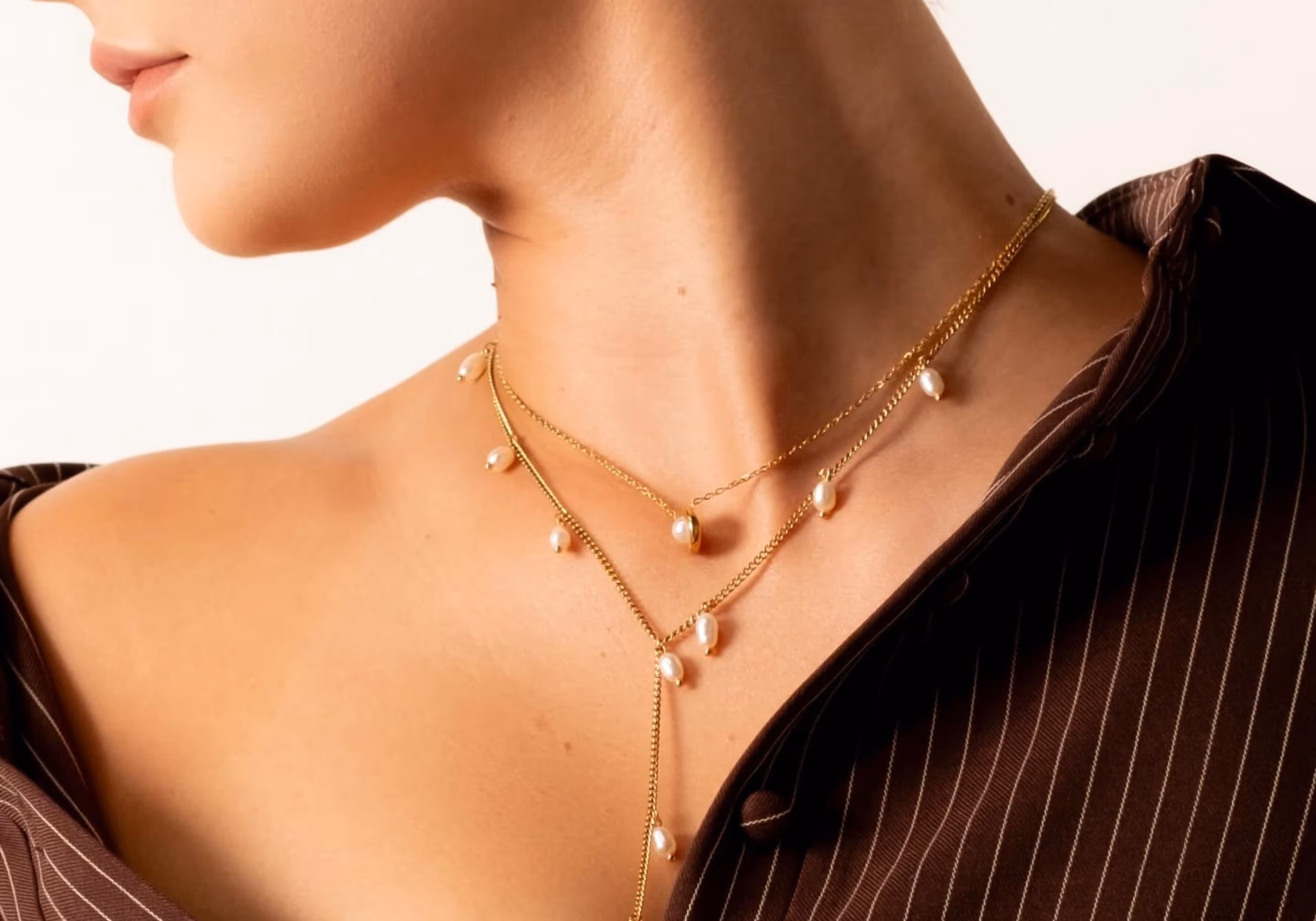 Close-up of a woman wearing layered gold necklaces with small pearls against a brown pinstripe shirt.