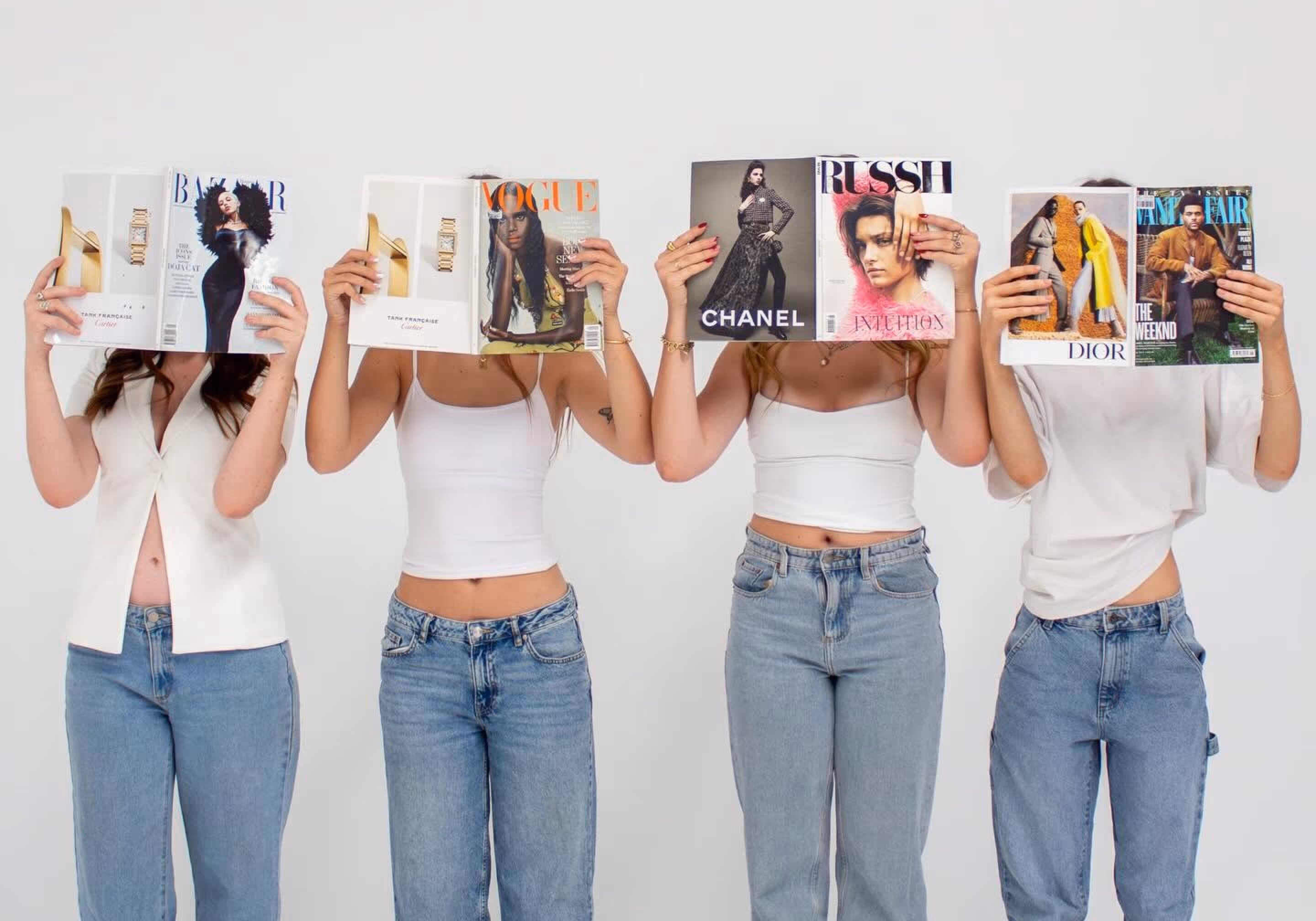 Four women wearing white tops and jeans hold fashion magazines in front of their faces against a plain white background.