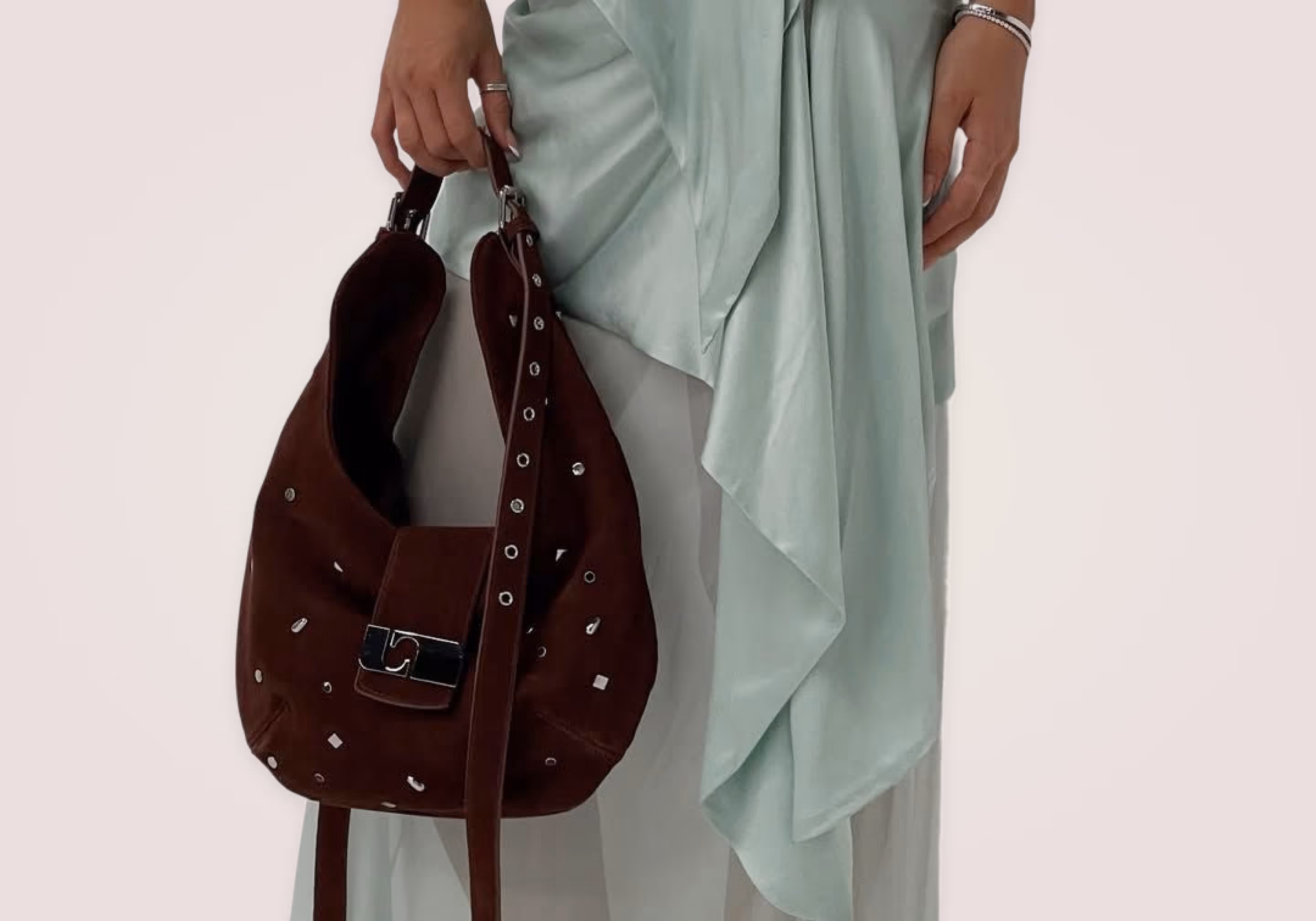 Woman holding a dark brown suede handbag with silver studs, wearing a light green flowing dress.