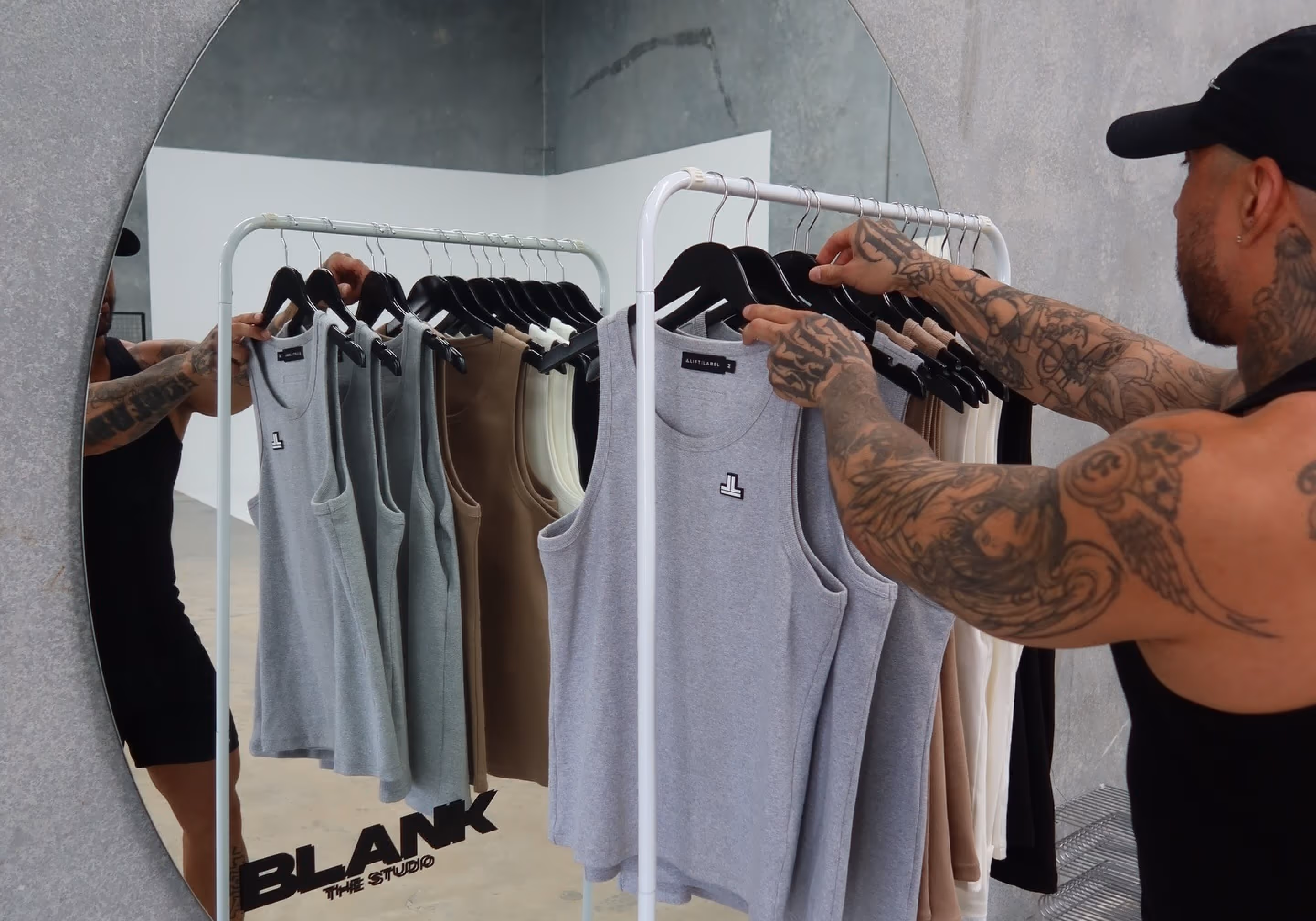 Tattooed man in a black cap adjusting sleeveless shirts on a white clothing rack in front of a large round mirror.