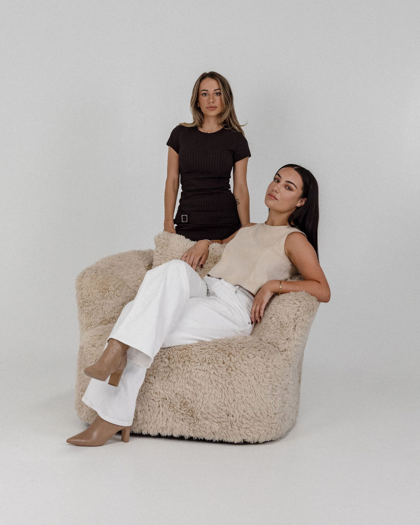 Two women posing, one sitting on a fluffy beige chair wearing cream and white, the other standing in black dress.