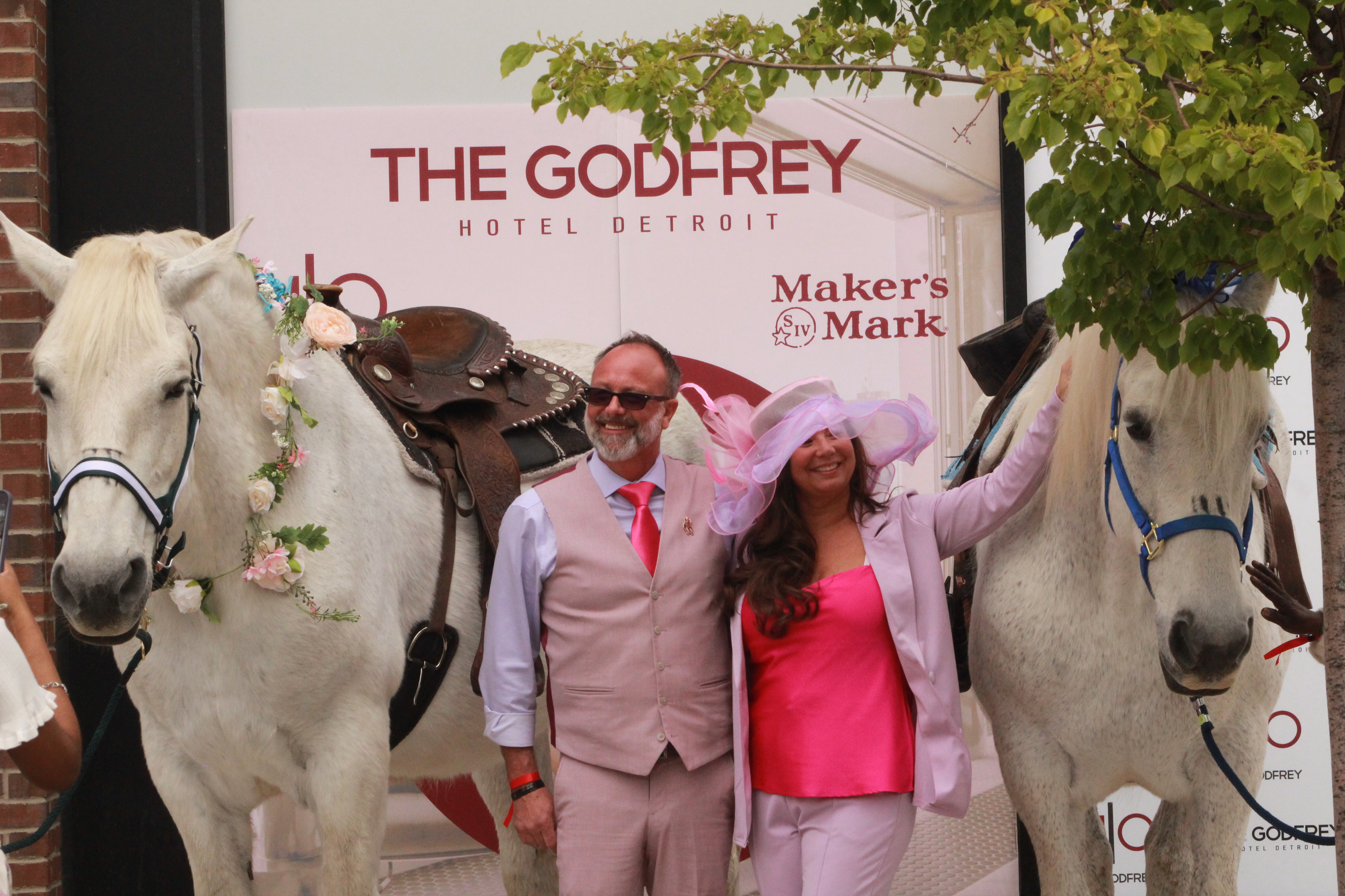  In front of the Godfrey Hotel Detroit, matching with pink suites, a husband (left) and wife (right) stand in between two grey, female Percherons (horses)