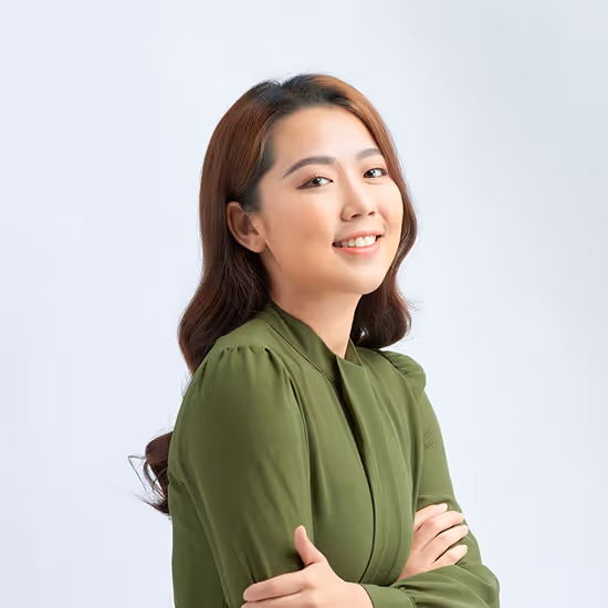 Smiling young woman with long dark hair wearing a green blouse, posing with folded arms against a light background.