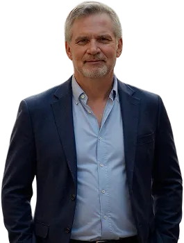 Middle-aged man with gray hair and beard wearing a dark blazer and light blue shirt, standing and smiling slightly.