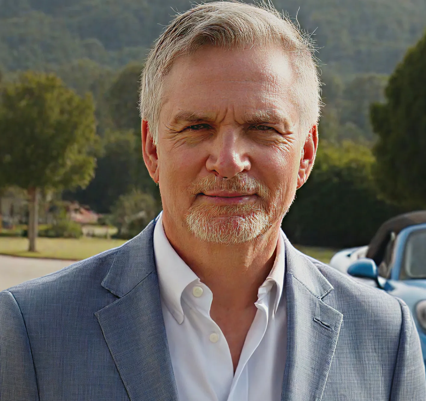 Mature man with gray hair and beard wearing a light blue blazer and white shirt outdoors with trees and a blue car in the background.