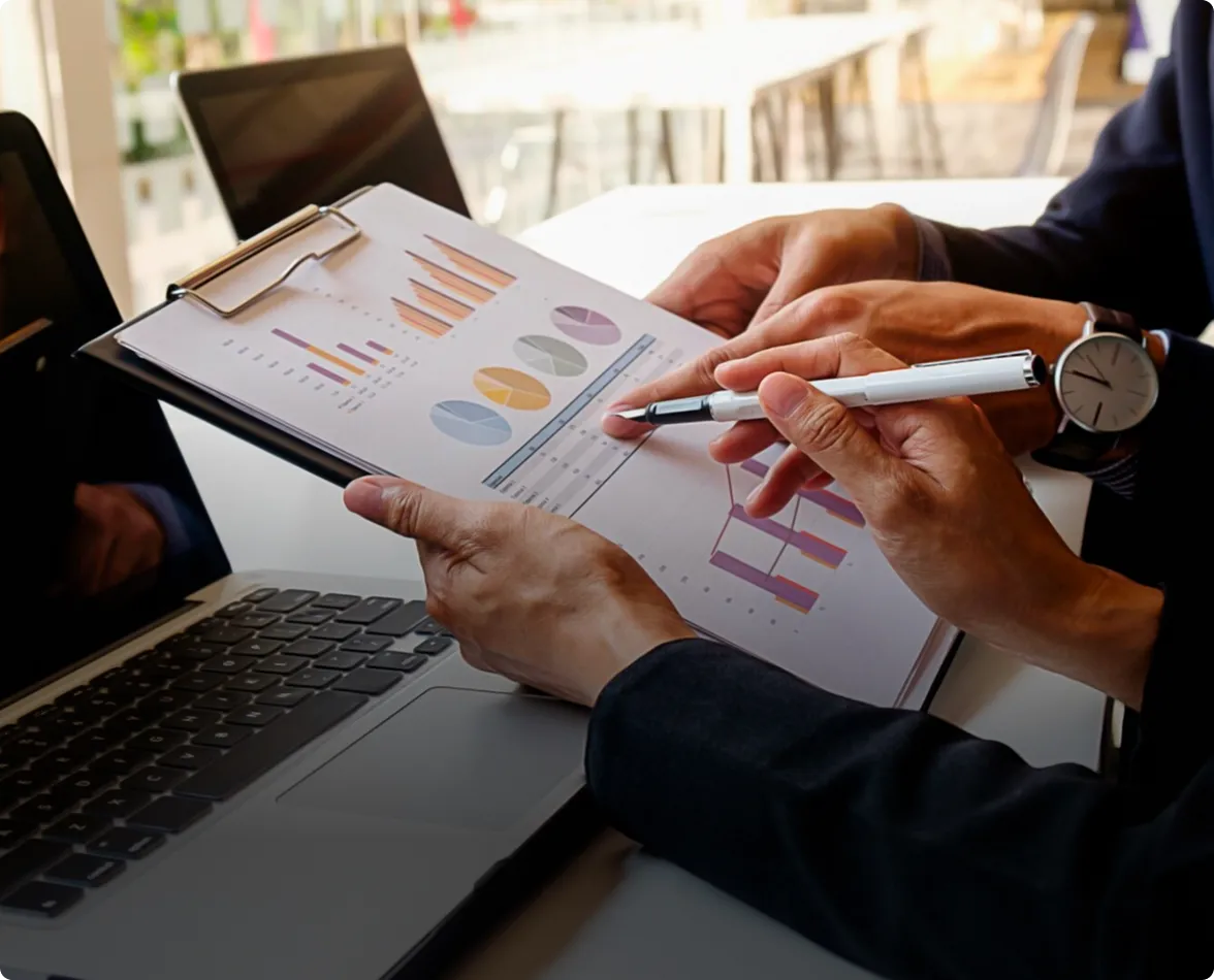 Two people reviewing charts and graphs on a clipboard beside a laptop during a business meeting.