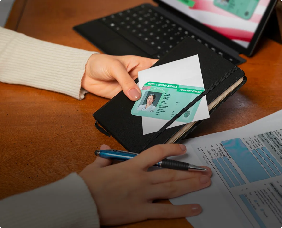 Person holding a black notebook with a U.S. permanent resident card and an envelope on top, next to a pen and a partially visible form on a wooden desk.