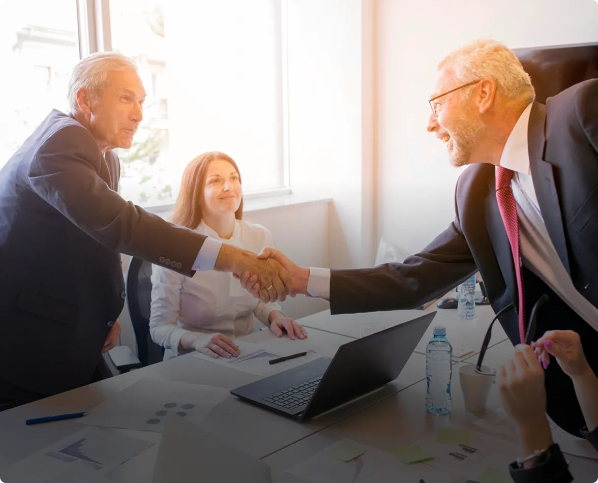 Two older men in suits shaking hands across a conference table while a woman in white watches and smiles.