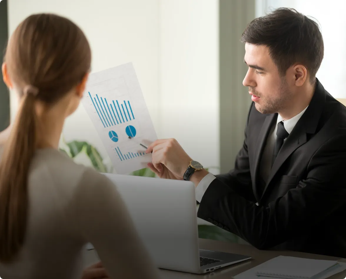 Businessman in a suit explaining charts and graphs on a paper to a woman in an office setting.
