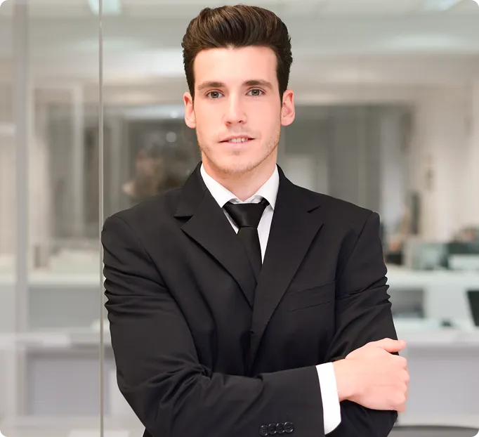 Young man in black suit and tie with arms crossed, standing in a modern office environment.