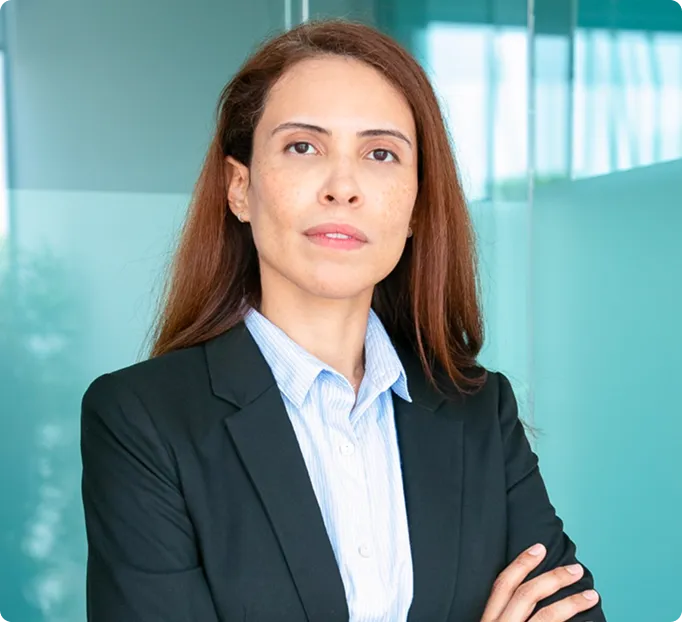 Professional woman with long brown hair wearing a black blazer and light blue shirt, standing with arms crossed in an office.