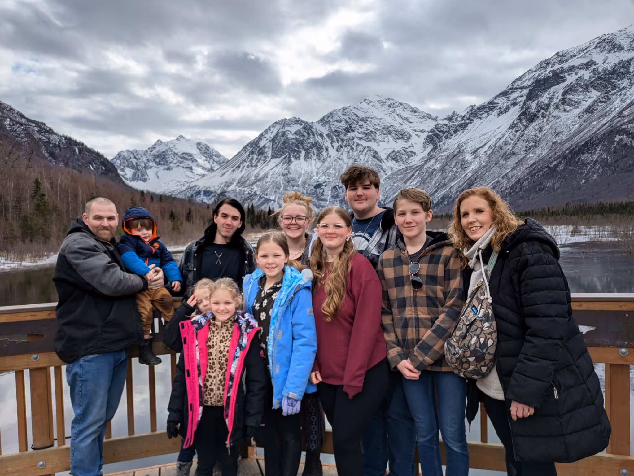 Group of eleven people posing on wooden deck with snowy mountains and cloudy sky in background.