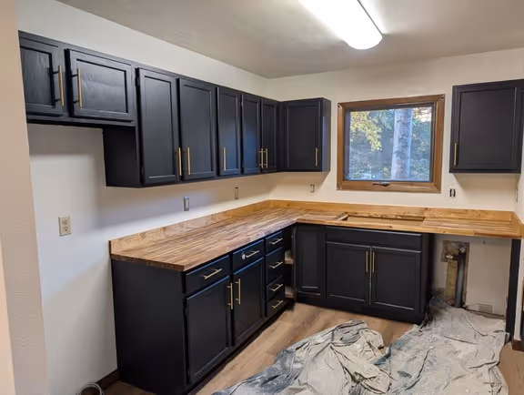 L-shaped kitchen with black cabinets, wooden countertops, and a window showing outdoor trees.