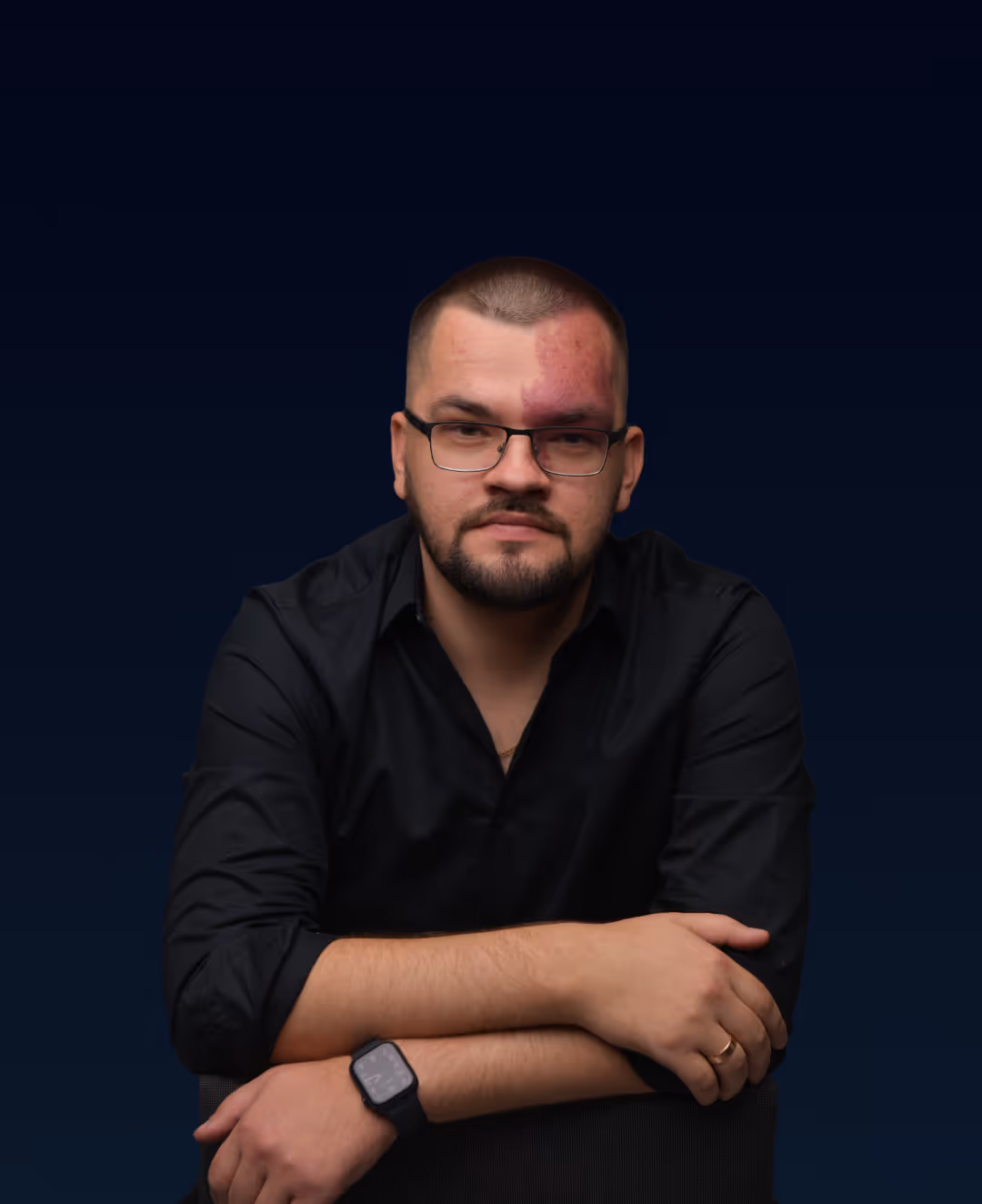 Man with glasses and a beard wearing a black shirt, seated with arms crossed, against a dark blue background.