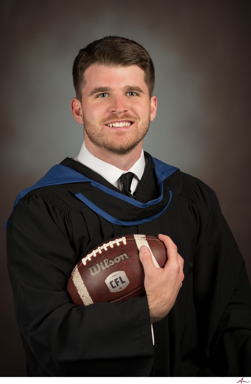 UBC graduate in black gown and blue hood holding a CFL football, studio grad portrait, Halifax