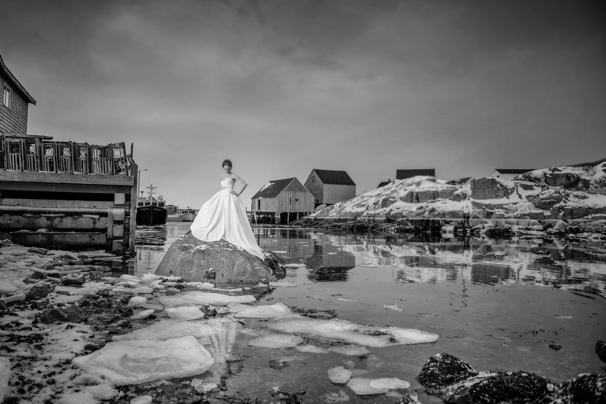Bride standing on a rock at Peggy's Cove in winter with ice in the harbour, black and white