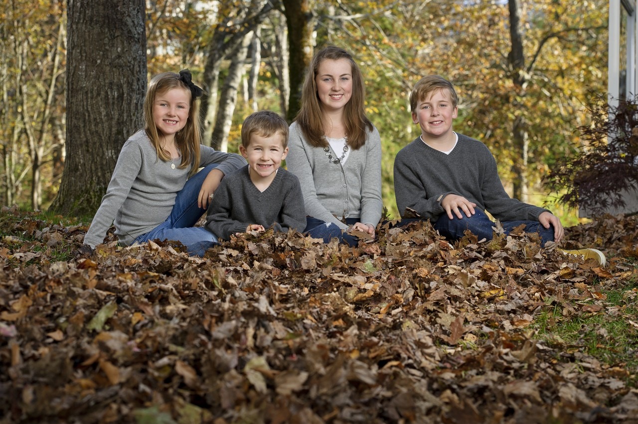 Four siblings in grey sweaters sitting in fallen oak leaves on a sunny fall day in Bedford