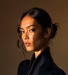 Portrait of a woman with dark hair styled loosely, wearing a black outfit, against a dark brown background.