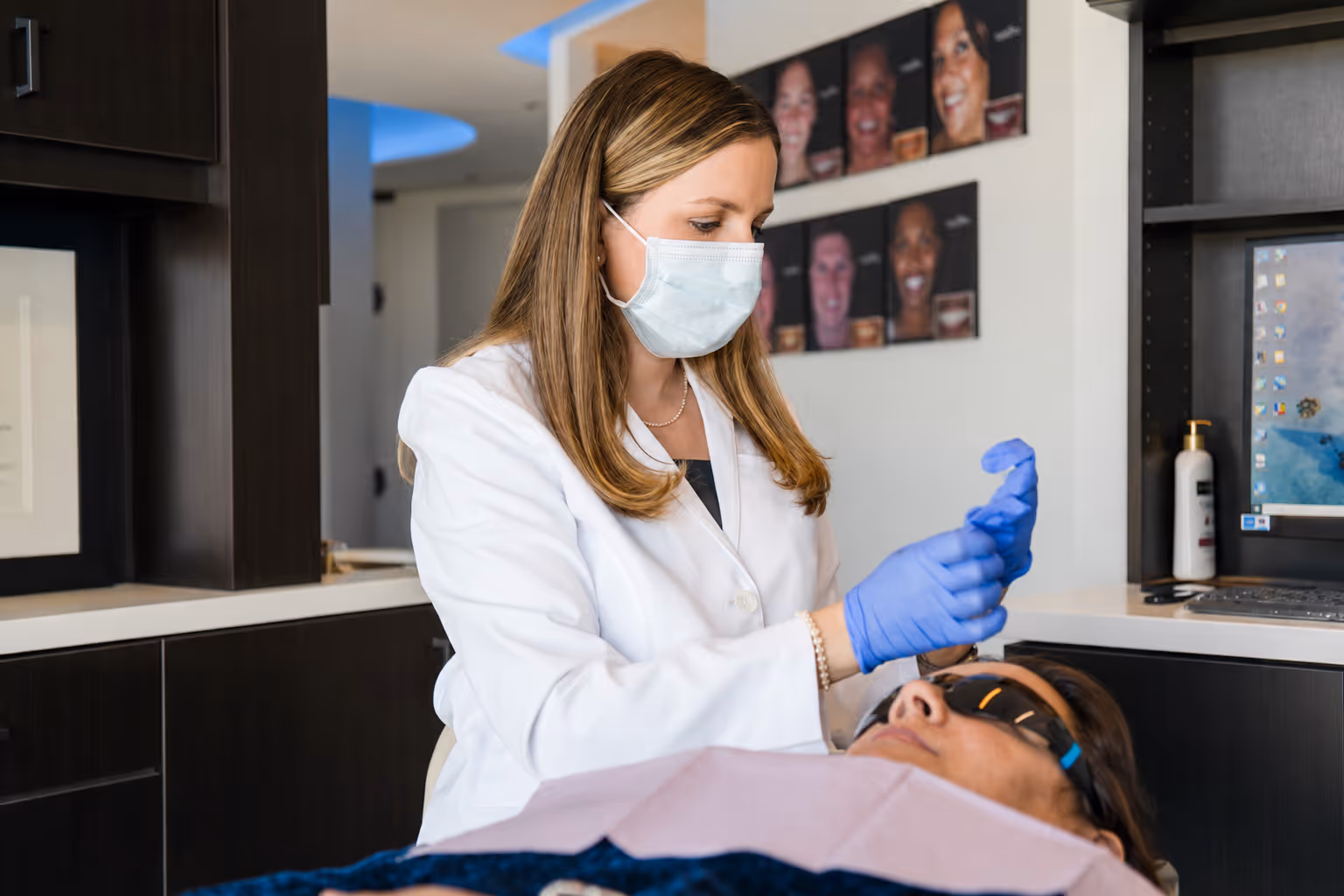 Dentist in white coat and mask putting on blue gloves over a patient wearing protective glasses.