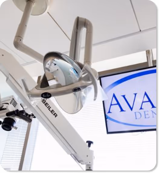 Dental operatory light and equipment arm next to a monitor showing partial text 'AVA DENT'.