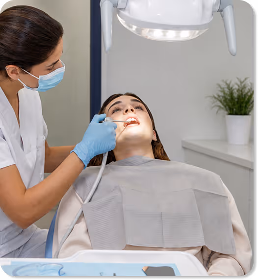 Dentist wearing mask and gloves examines a female patient's teeth under dental light.