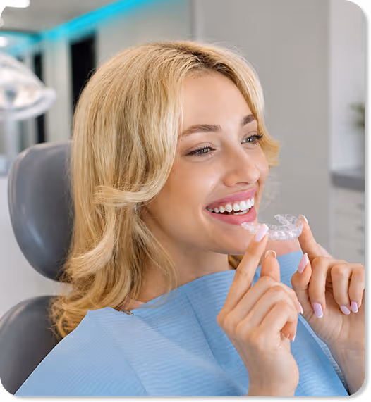 Smiling woman in dental chair holding a clear orthodontic aligner tray near her mouth.
