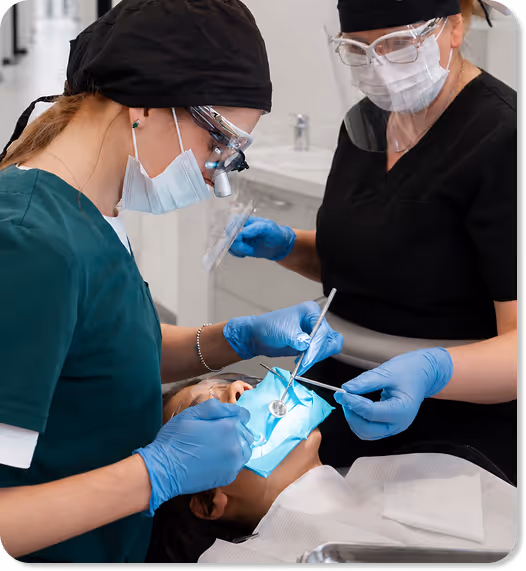 Two dental professionals wearing masks and gloves performing dental work on a patient reclining in a chair.