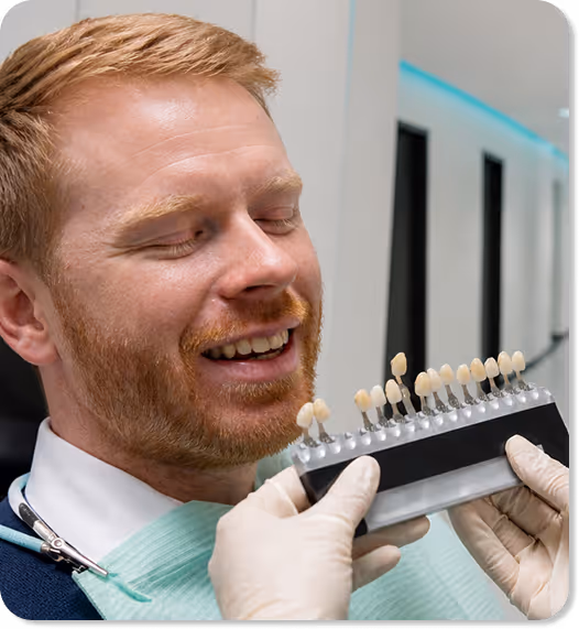 Man with red hair smiling as dentist holds a tooth shade guide near his mouth.