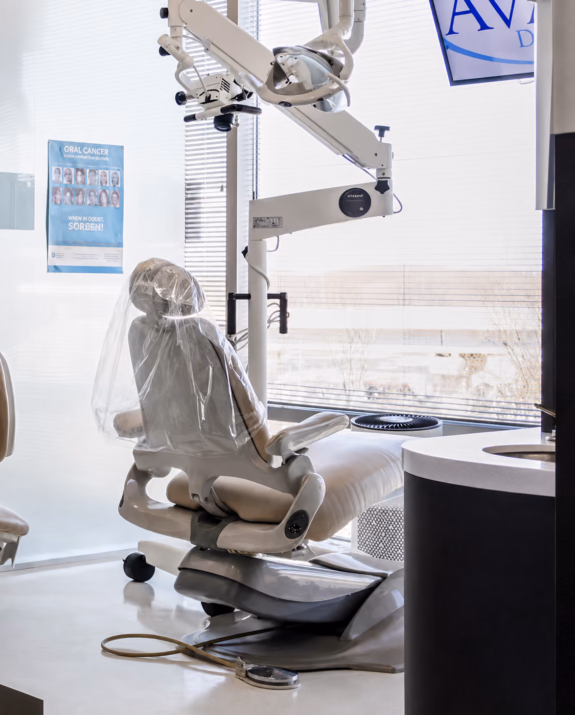 Modern dental chair with protective plastic cover and overhead dental equipment near window blinds.