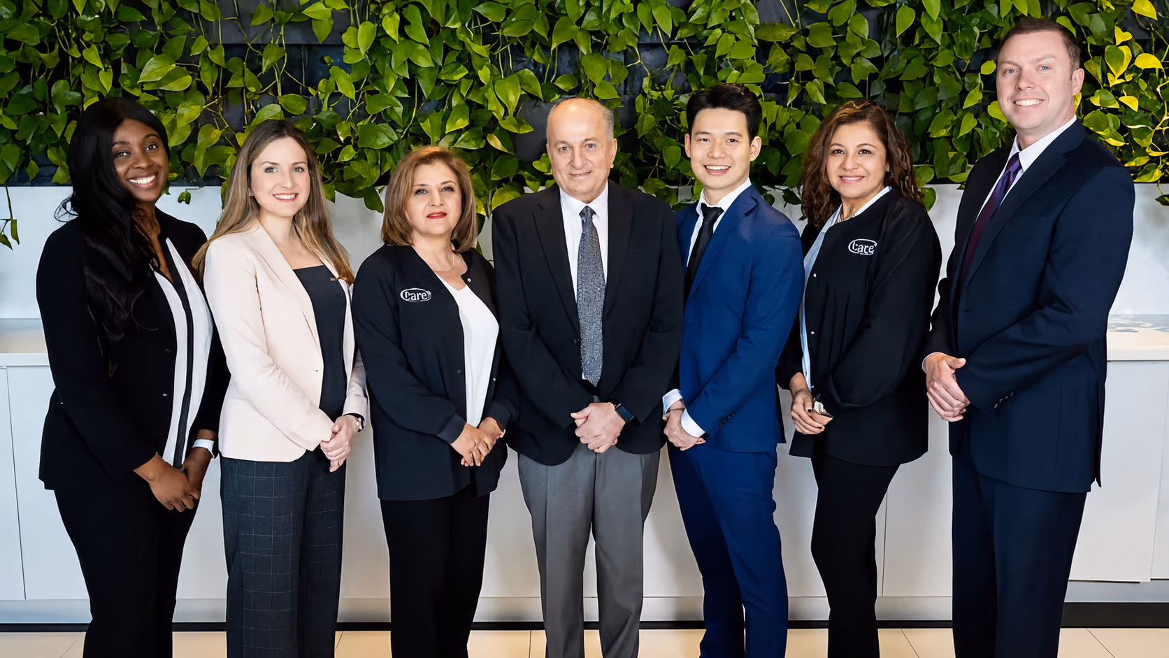 Seven diverse professionals standing side by side, smiling against a green leafy wall background.