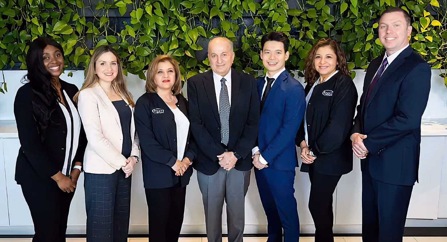Seven diverse professionals standing side by side, smiling against a green leafy wall background.