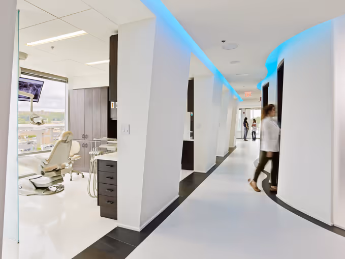 Modern dental office hallway with white floors, blue ceiling lights, and a dentist walking past treatment rooms.
