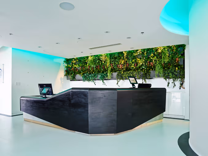 Modern black reception desk with two monitors and a green plant wall behind it in a bright office lobby.