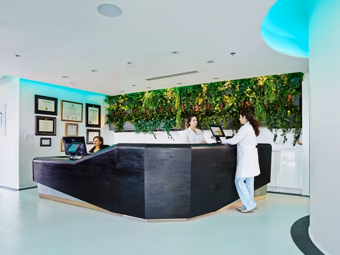 Modern medical reception area with staff at black counter and green plant wall behind.