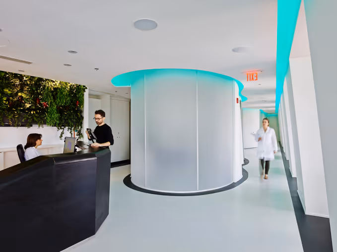 Modern medical reception area with a receptionist, a visitor, and a doctor walking down a hallway.