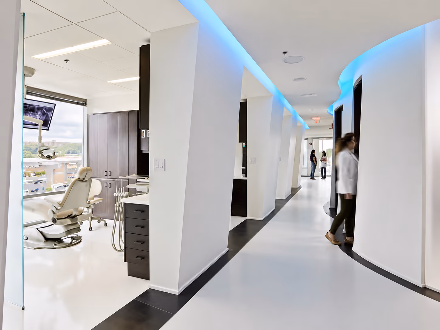 Modern dental clinic hallway with treatment rooms and dental chair by a window.