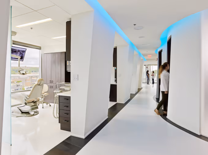 Modern dental clinic hallway with treatment rooms and dental chair by a window.