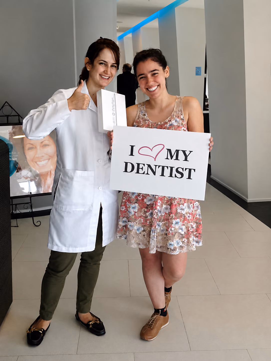 Smiling dentist and patient holding a sign that says 'I heart my dentist' inside a clinic.