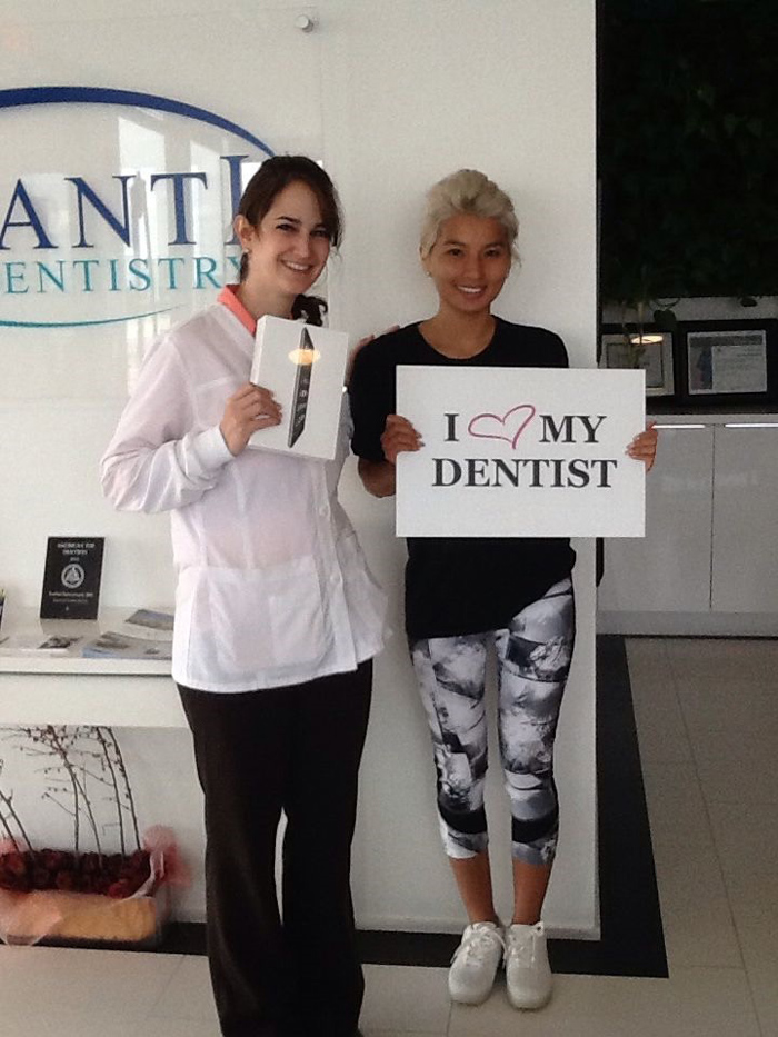 Two women smiling at dental office; one holds an iPad box, the other a sign saying I love my dentist.