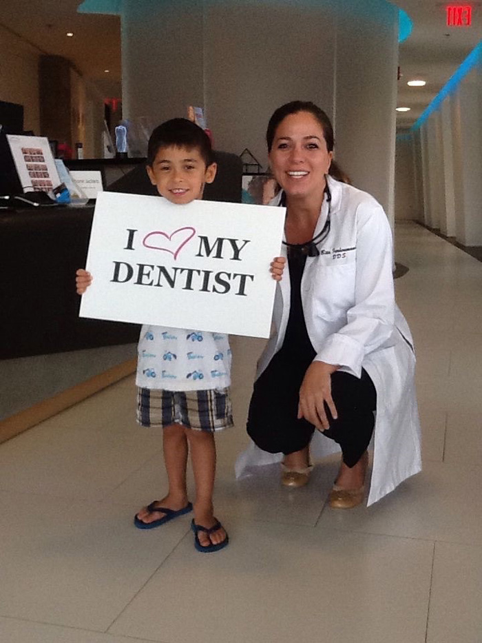 Smiling boy holding a sign saying 'I ♥ MY DENTIST' next to a female dentist in a white coat.