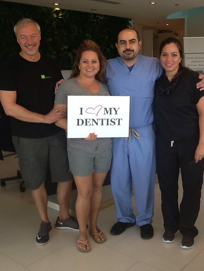 Four people smiling, one holding a sign reading 'I ♥ MY DENTIST' inside a dental office.