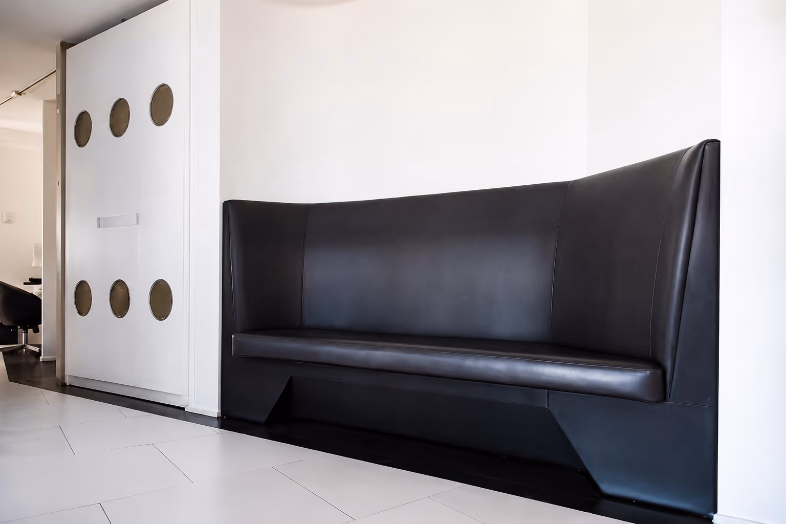 Modern black leather bench in a white, minimalist room with sliding white doors featuring circular cutouts.