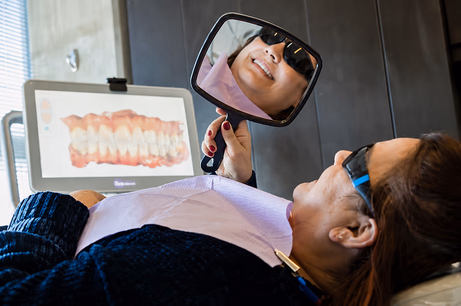 Patient wearing sunglasses looking at her teeth in a hand mirror in a dental clinic.