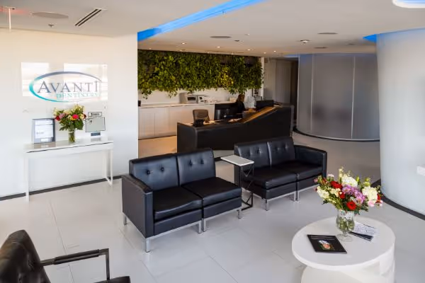Modern office lobby with black leather sofas, white tables, flowers, and a receptionist desk with green wall.