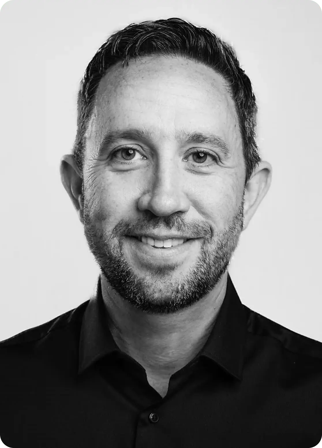 Smiling man with short dark hair and beard wearing a black button-up shirt on white background.