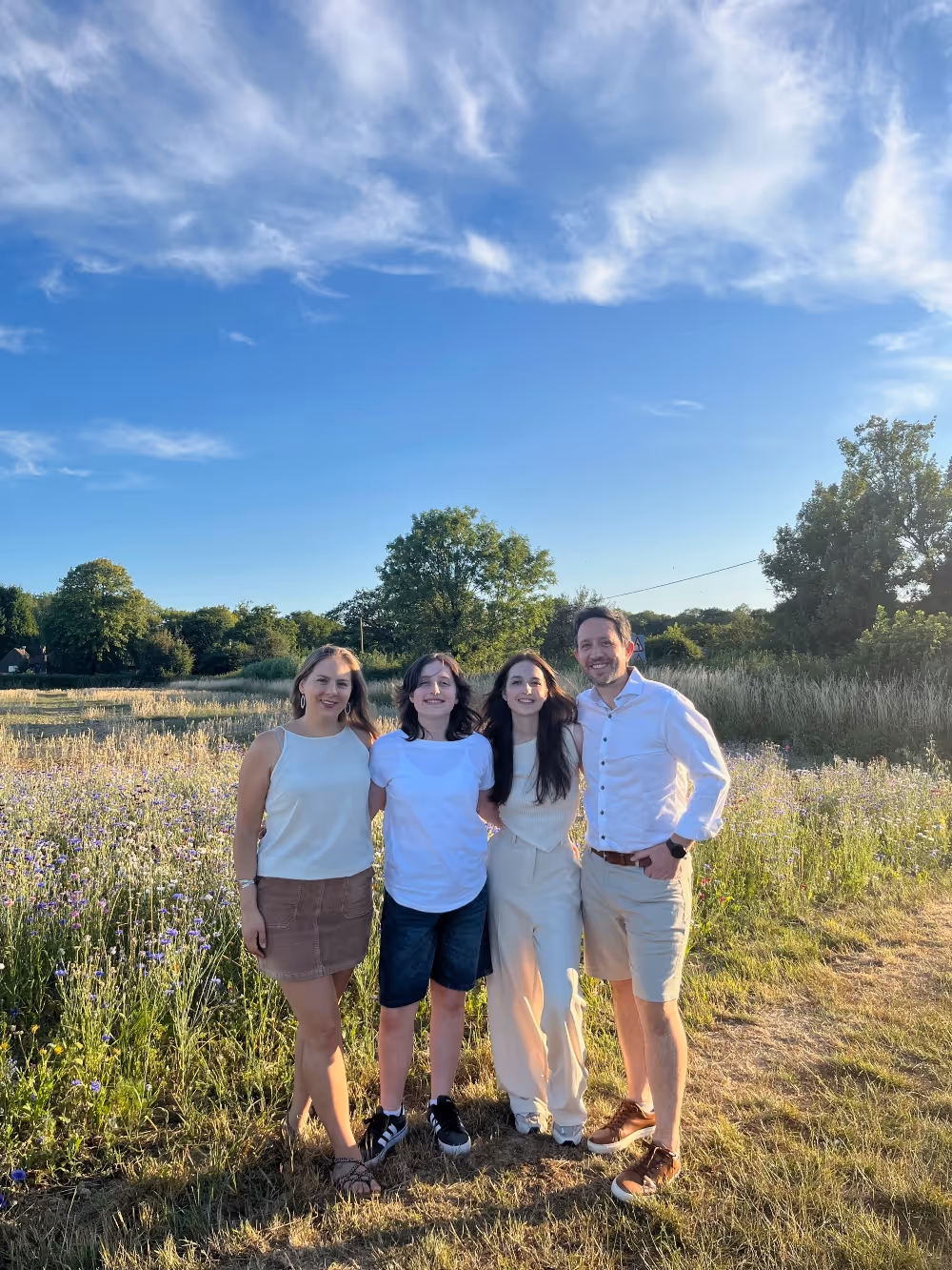 Four people standing together smiling in a grassy field with trees under a blue sky.
