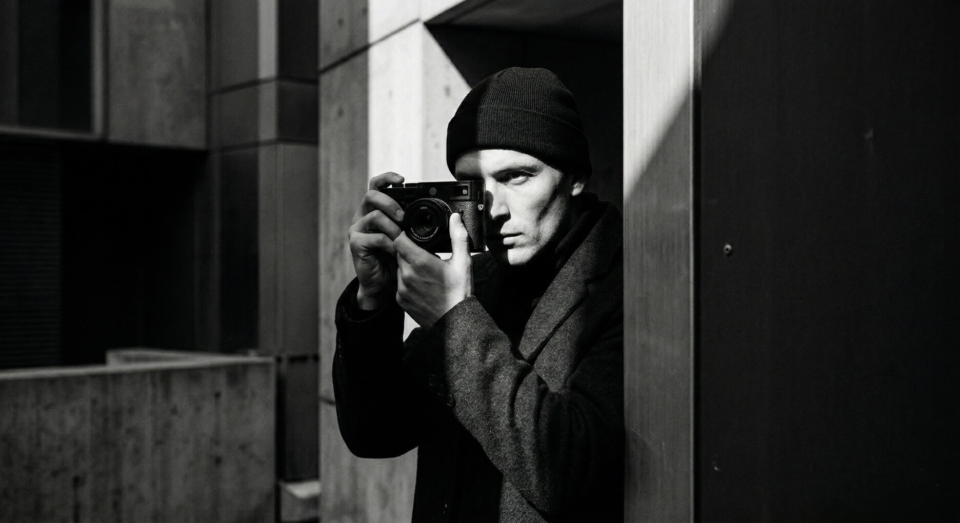Man in beanie and coat holding a camera to his eye in a high-contrast black and white urban scene.