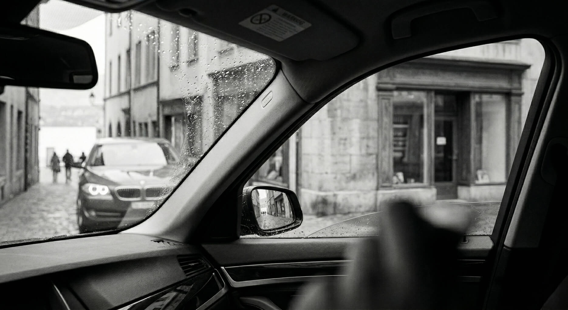 View through a car window on a rainy street with a black car and people walking in the distance.