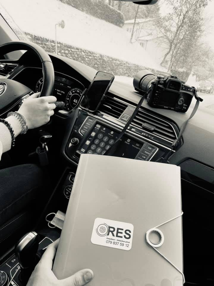 Inside car showing steering wheel held by driver, dashboard, camera on dashboard, and hand holding ORES folder.