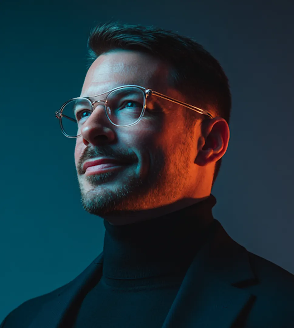 Man with glasses and a beard wearing a black turtleneck and jacket looking thoughtfully to the side.